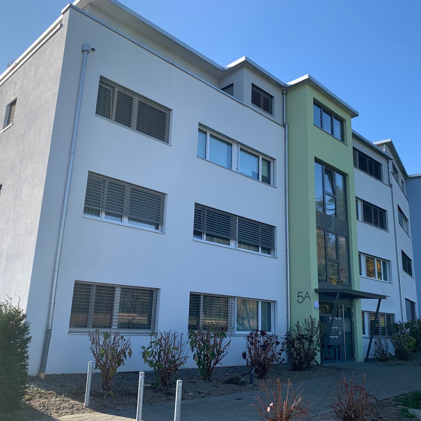Mehrfamilienhaus mit weisser Fassade und grün abgesetztem Treppenhaus, daneben der Hauseingang 5A