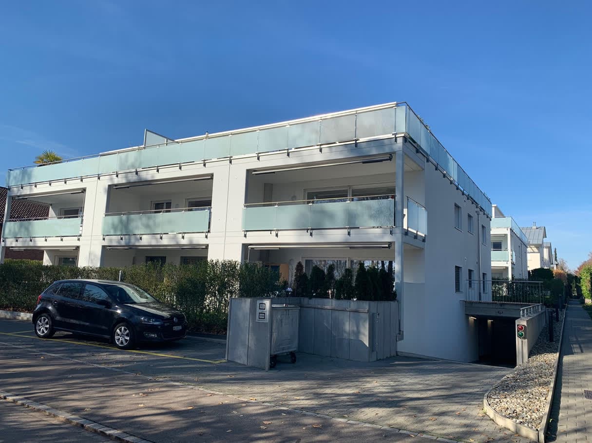 Mehrfamilienhaus mit Glasbrüstungen, davor Besucherparkplätze und die Garageneinfahrt