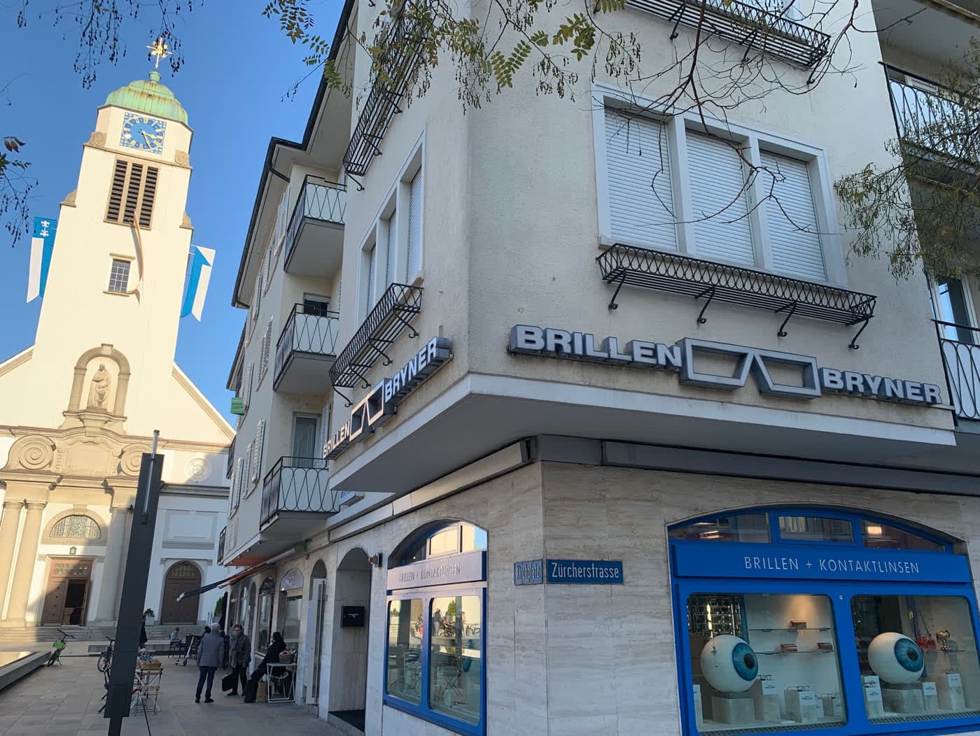 Eckhaus mit Ladenlokal im Erdgeschoss, dahinter der Turm der Dorfkirche