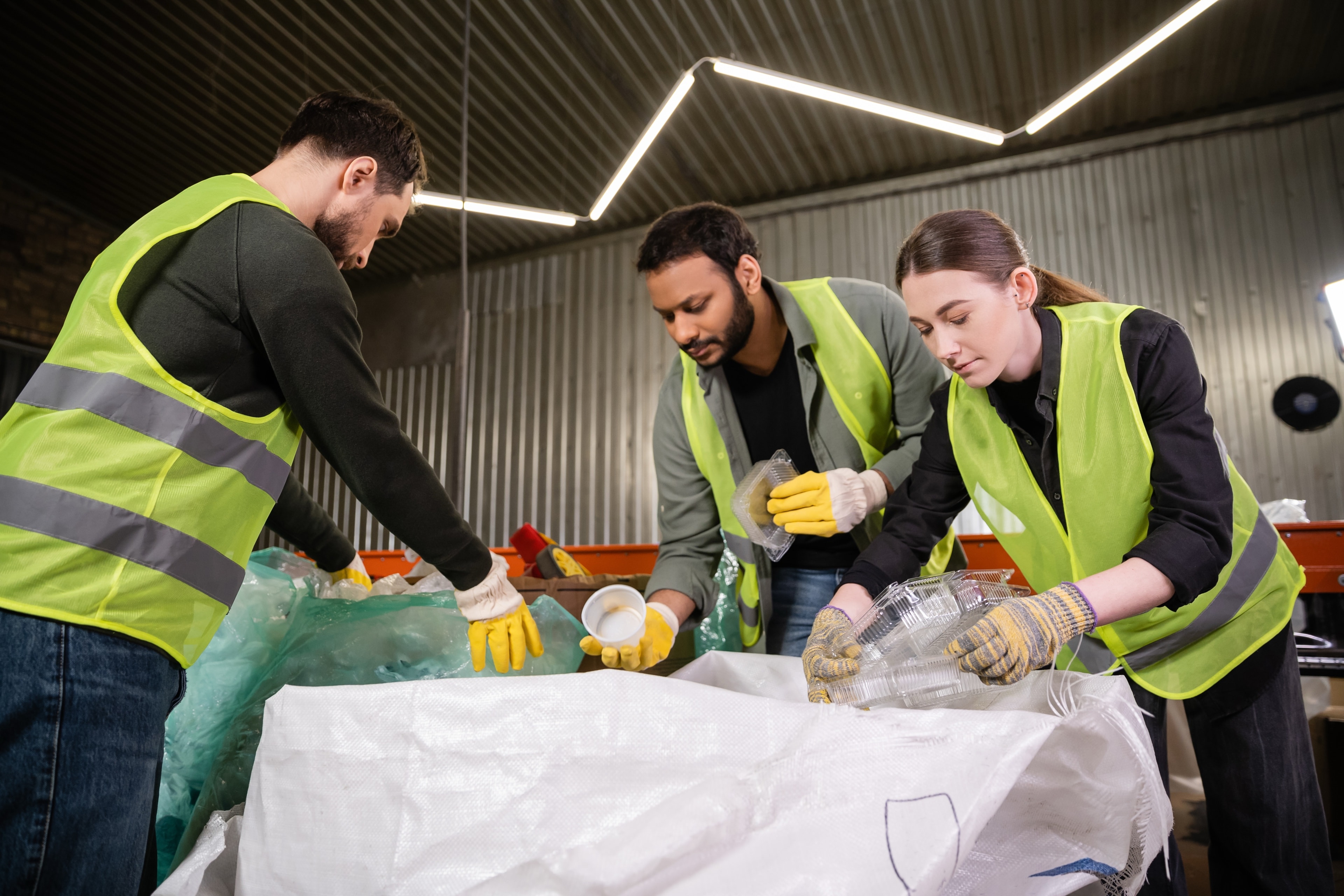 Drei Mitarbeitende in Warnwesten sortieren Kunststoffverpackungen an einer Sammelstelle in Wertstoffsäcke