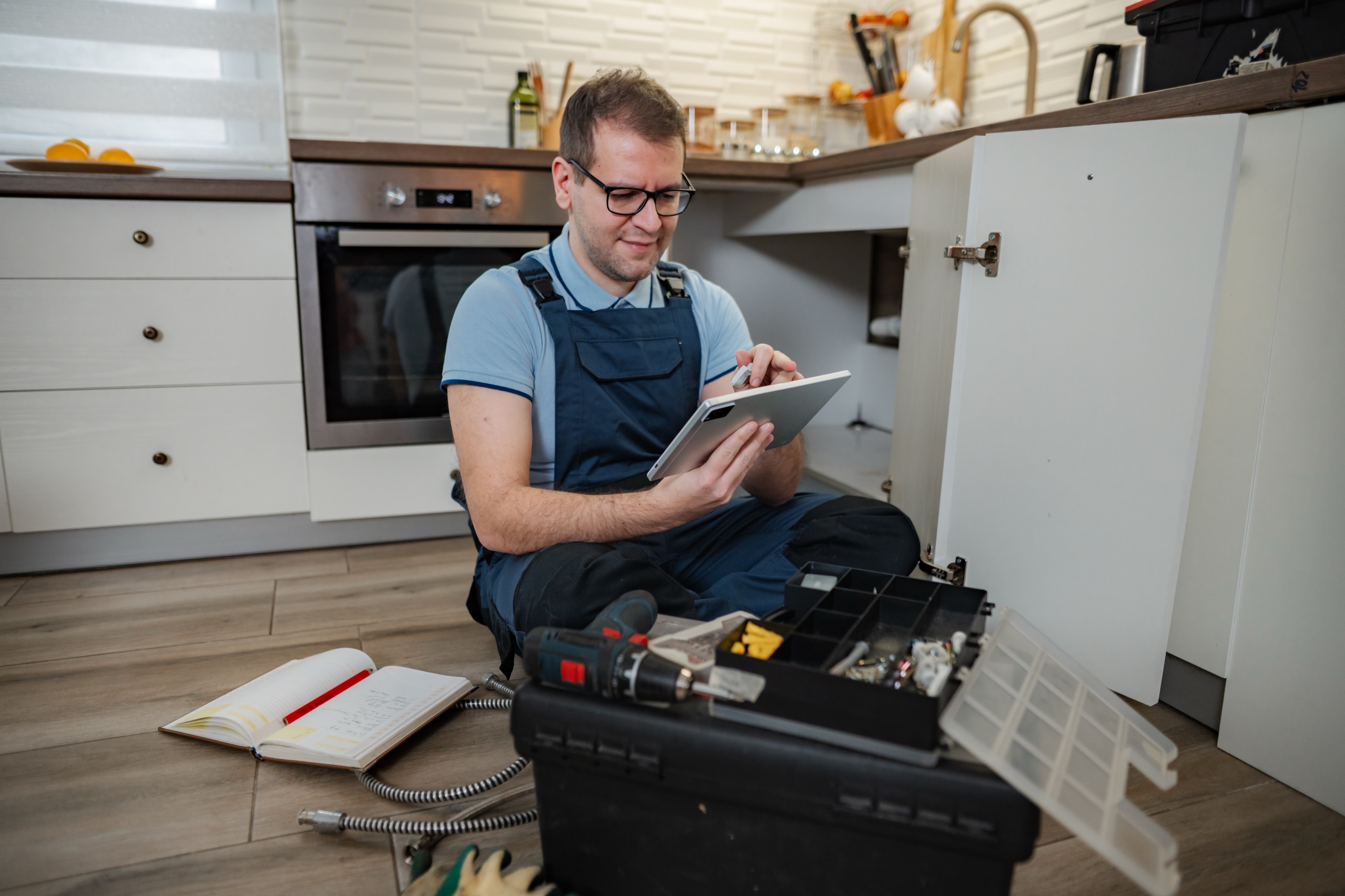 Ein Hauswart sitzt mit Werkzeugkoffer und Tablet vor einem geöffneten Küchenunterschrank und hält eine Kleinreparatur fest