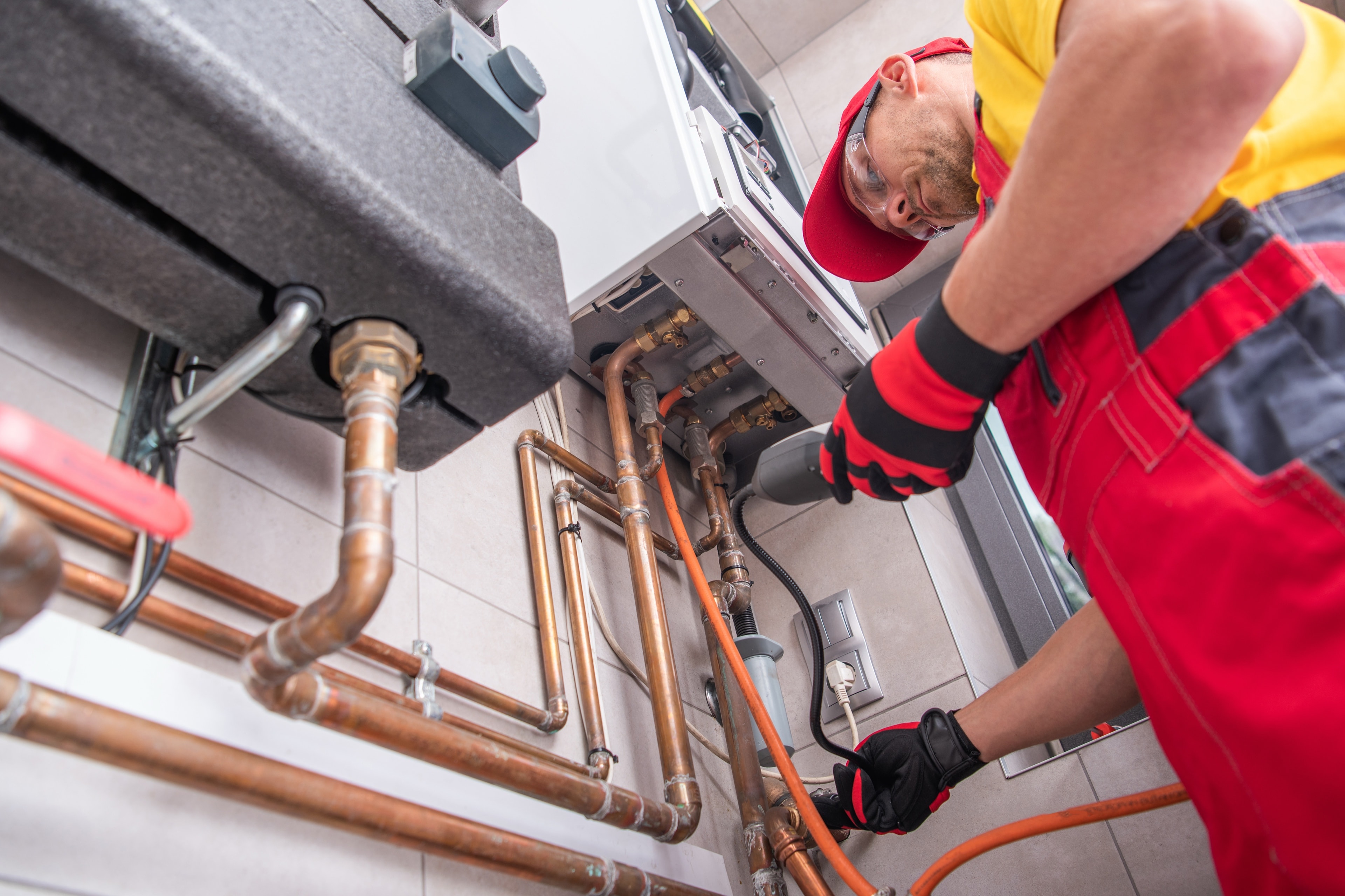 Ein Haustechniker in roter Arbeitskleidung prüft die Kupferleitungen einer Gastherme in einem Technikraum